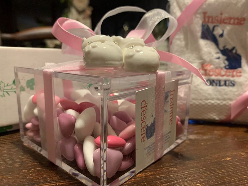 Una scatola regalo trasparente piena di caramelle rosa e bianche, sormontata da due mandorle ricoperte di caramelle bianche e un nastro rosa, su un tavolo disordinato, destinata alle donazioni onlus. Una scatola regalo trasparente piena di caramelle rosa e bianche, sormontata da due mandorle ricoperte di caramelle bianche e un nastro rosa, su un tavolo disordinato, destinata alle donazioni onlus.