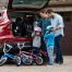 Due adulti e due bambini sono in piedi accanto al bagagliaio aperto di un SUV con biciclette e caschi per bambini, mentre si preparano per un giro in bicicletta nel vialetto e discutono suggerimenti su come gestire le coliche gassose durante le gite in famiglia.