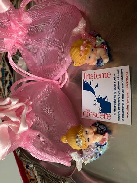 Su una superficie riflettente sono disposte due statuette di bambine, due sacchetti regalo in organza rosa e un biglietto da visita di "Insieme per Crescere".