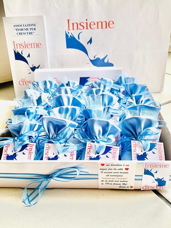 Un vassoio di piccoli sacchetti regalo blu e bianchi con il marchio "Insieme", disposti ordinatamente con un nastro, cartoline promozionali e un sacchetto di carta sullo sfondo.